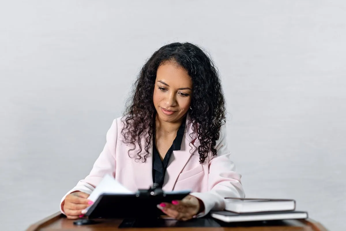 Woman in pale blazer turning pages of a black folder at a desk