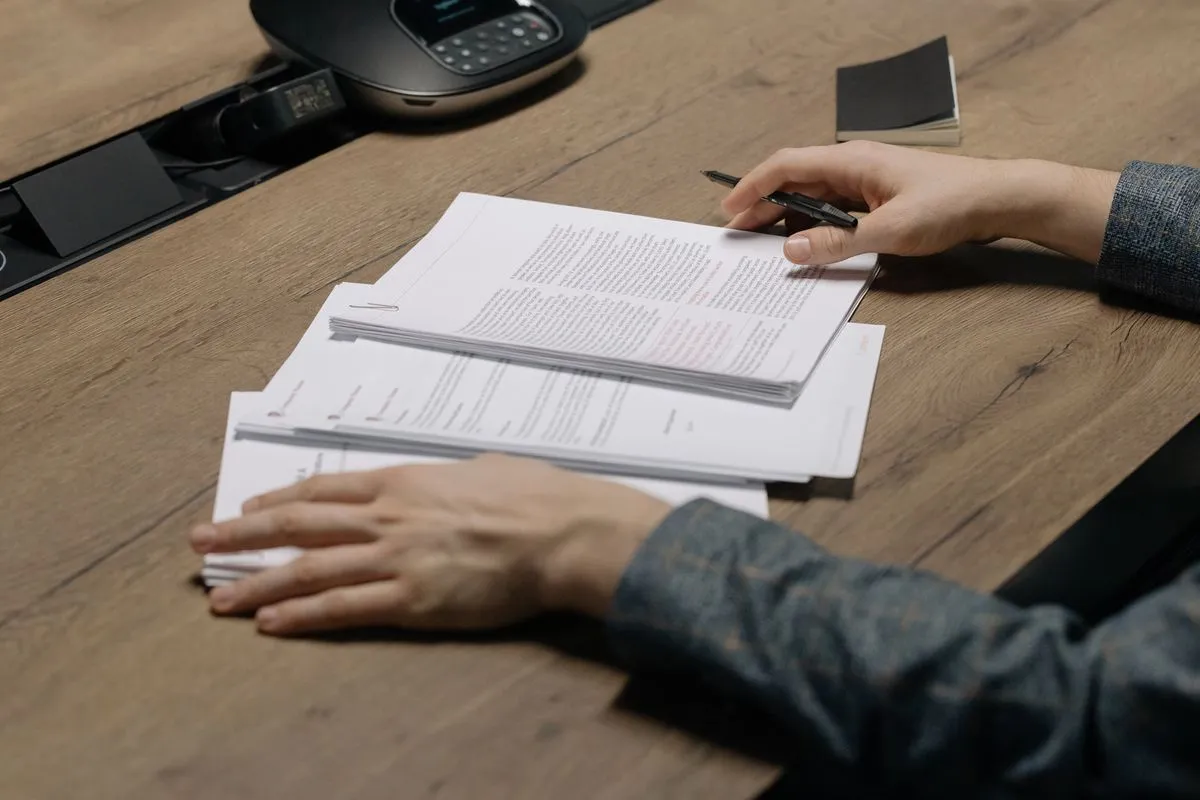 Hands over a stack of stapled printed pages, pen ready, wooden table