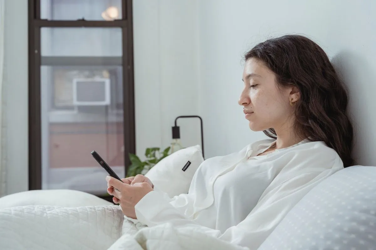 Woman in a white shirt sits up in bed reading her phone