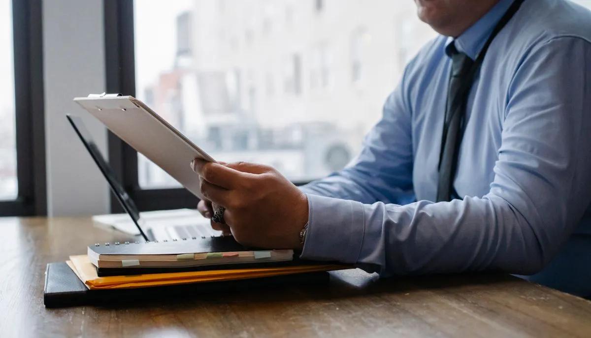 Seated man in shirt and tie reading a clipboard page beside an open laptop