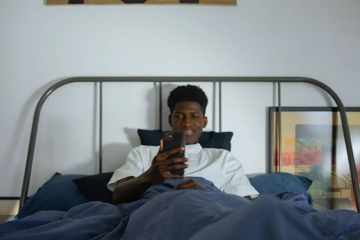 Man propped up in bed under a blue duvet, looking at his phone
