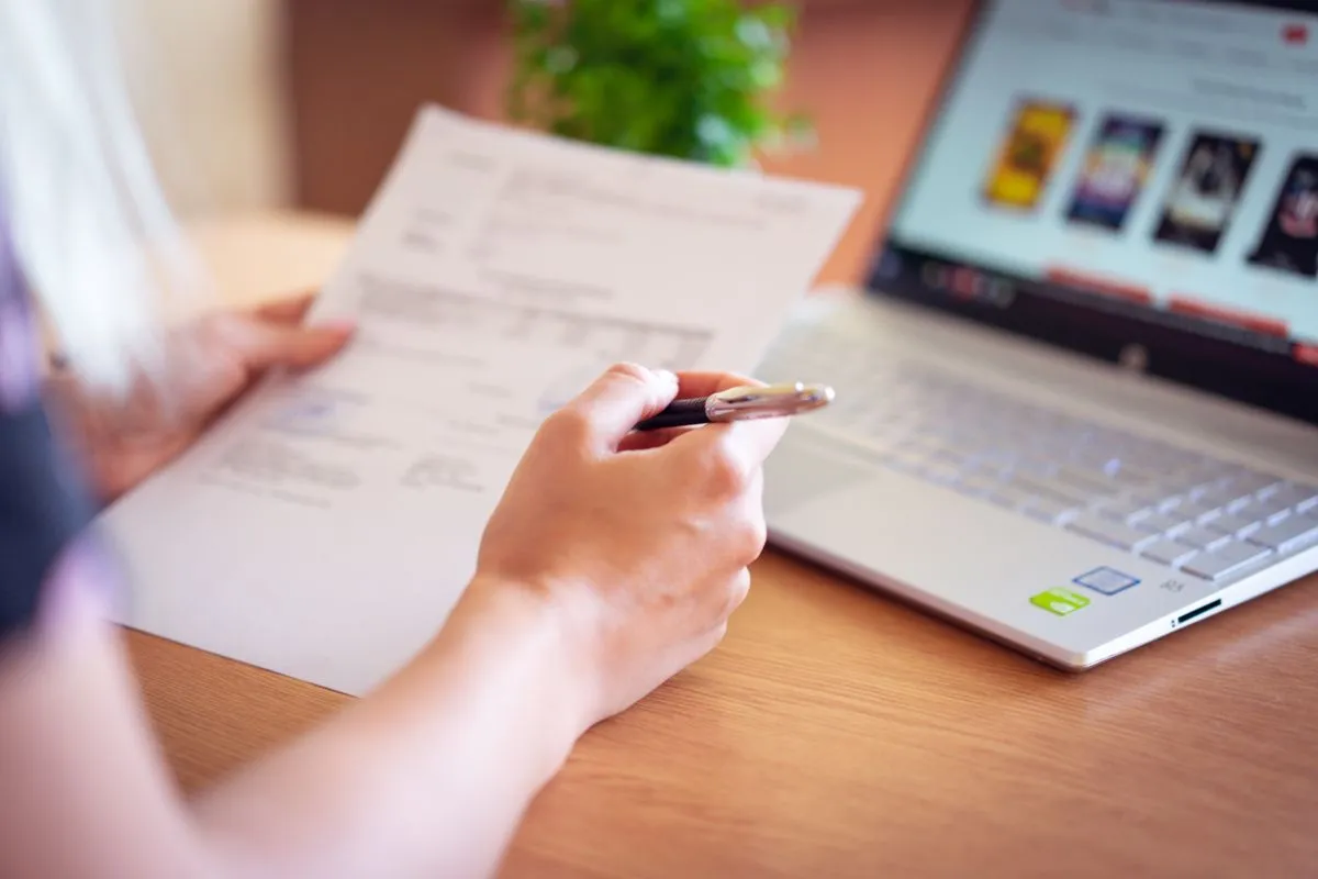 Hand holding a pen reviewing a printed form beside an open laptop