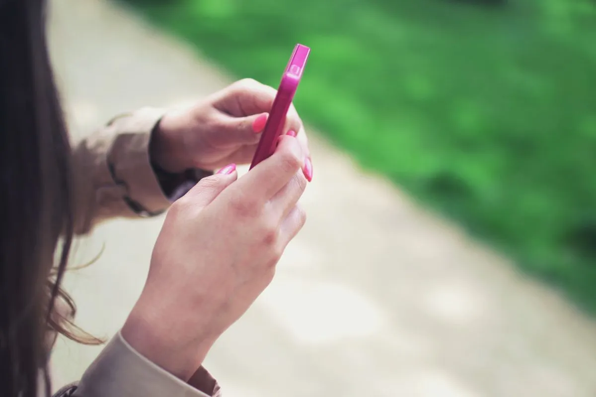 Woman's hands holding a slim pink phone on a sunlit path