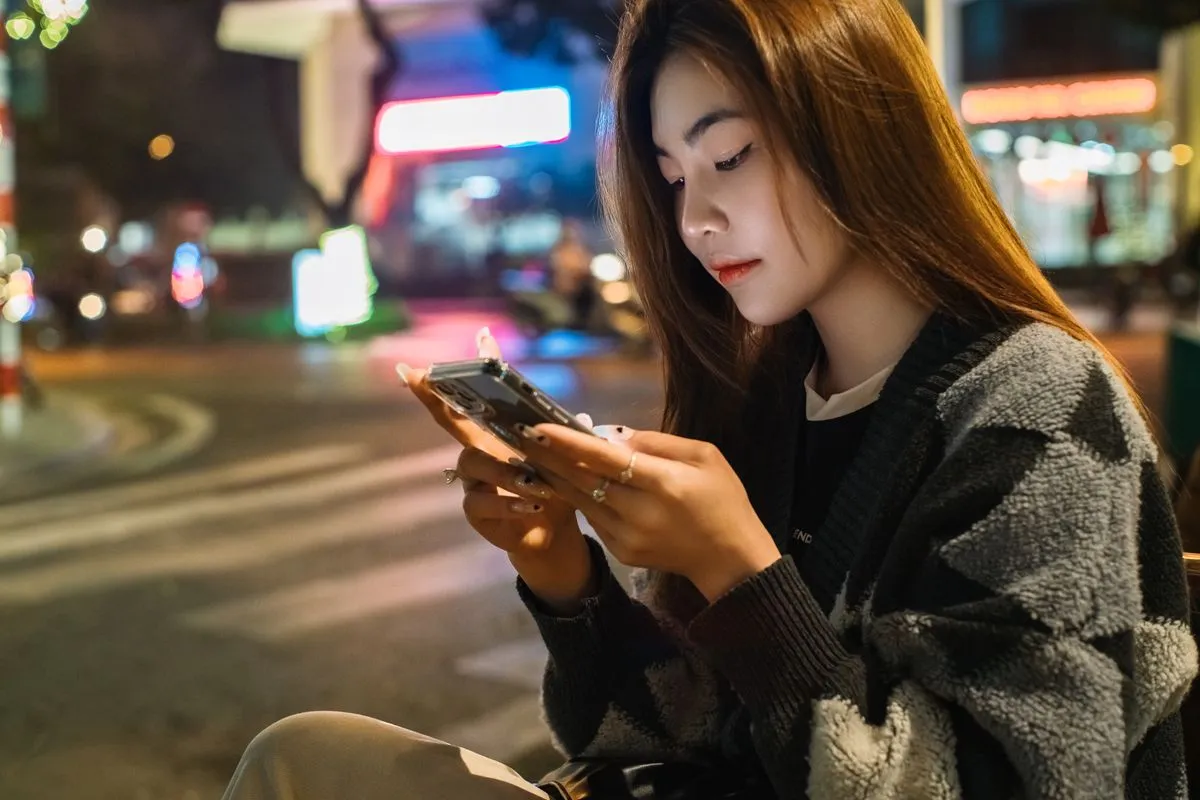 Young woman sitting on a neon-lit street at night, typing on her phone