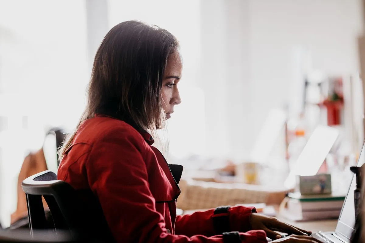 Woman in a red jacket typing at a laptop beside a bright window