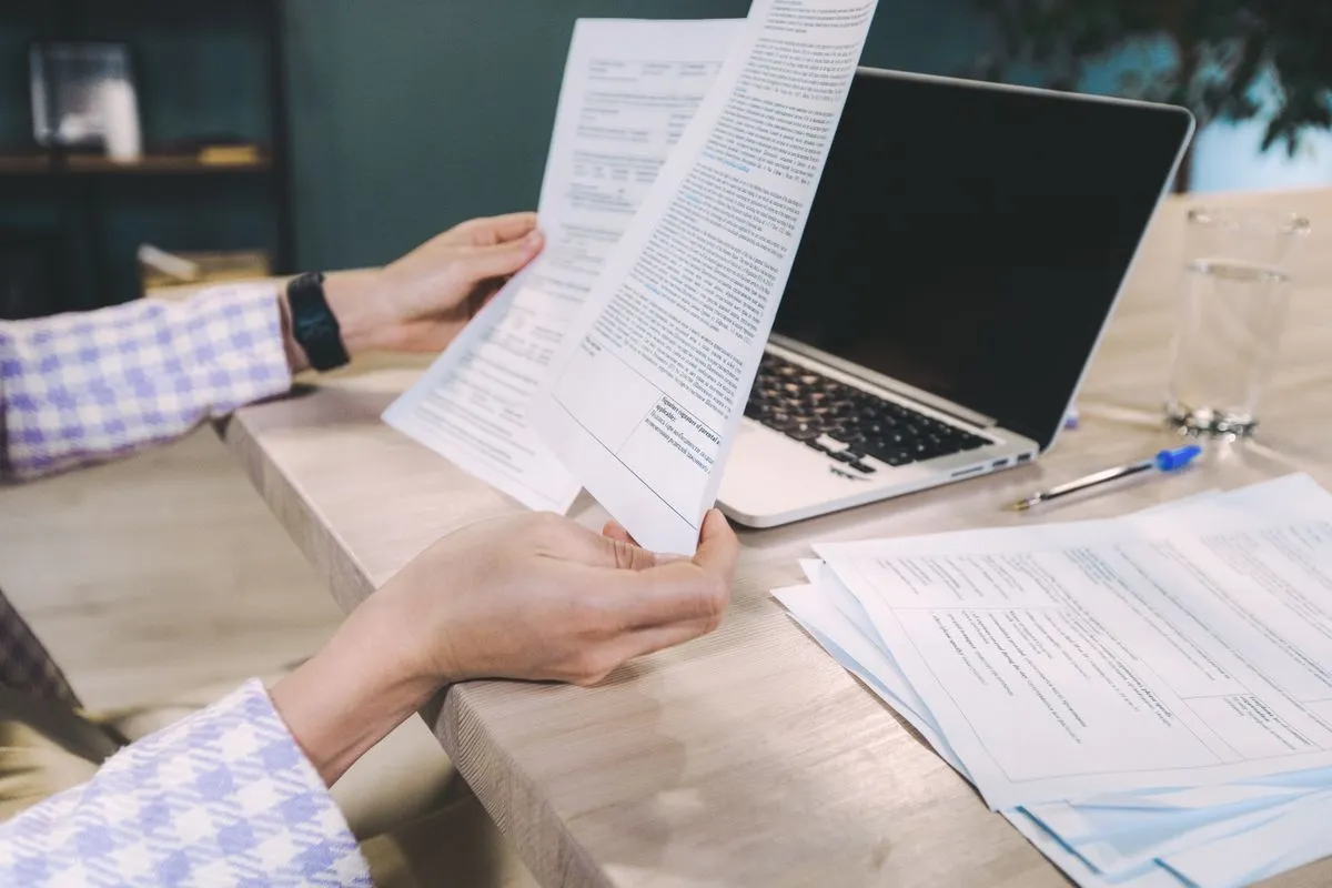 Hands lifting a printed page from a document stack beside a laptop
